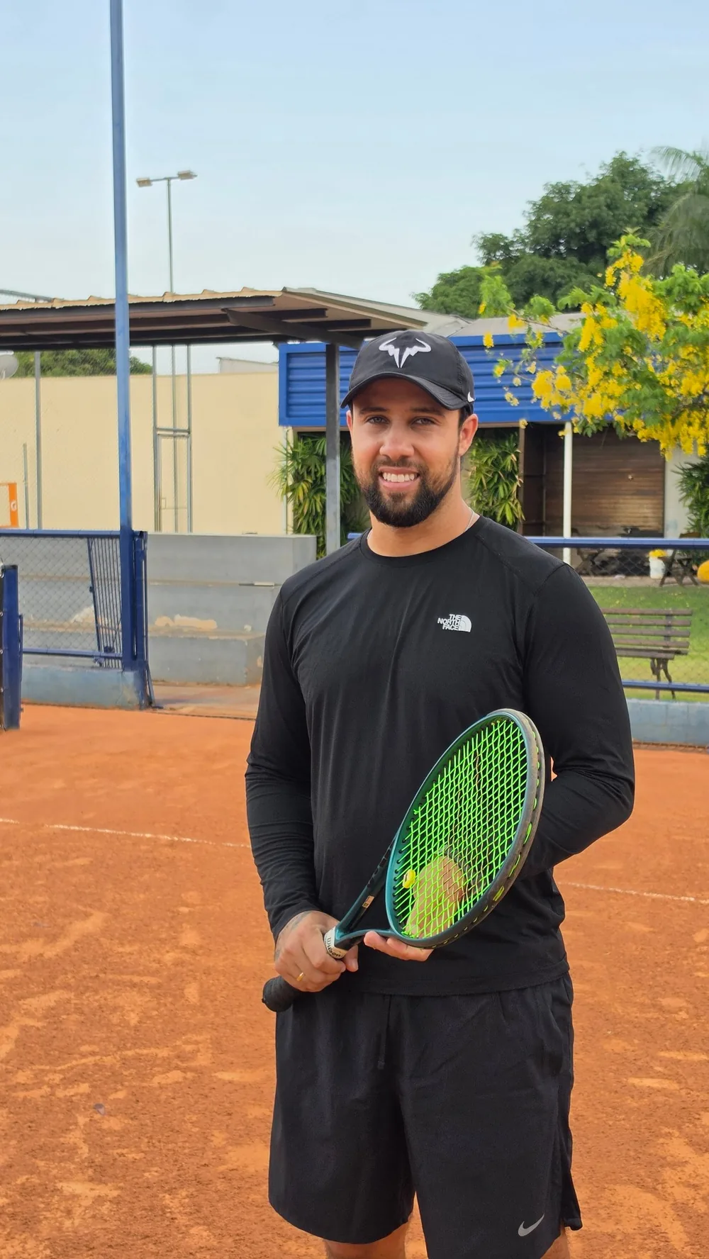 Lucas Marcolin, treinador de tênis, sorrindo em quadra de saibro com a raquete na mão